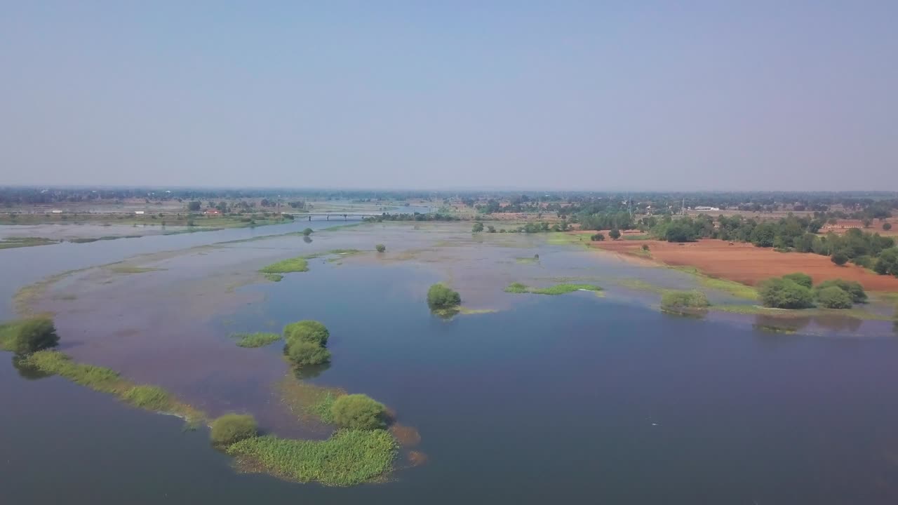 La zona umida del Gujarat presenta isole verdi sparse in acqua calma con campi agricoli e un'autostrada distante visibili. Una ripresa aerea fluida in avanti rivela un paesaggio rurale sotto un cielo limpido