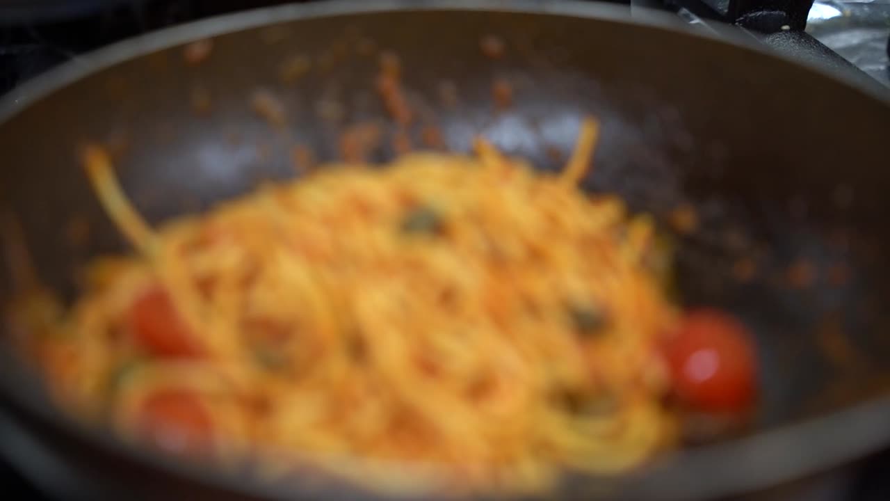 A detailed view of spaghetti mixed with a rich tomato sauce, garnished with cherry tomatoes and capers. The dish is presented in a pan, showcasing its vibrant colors and Mediterranean flavors.