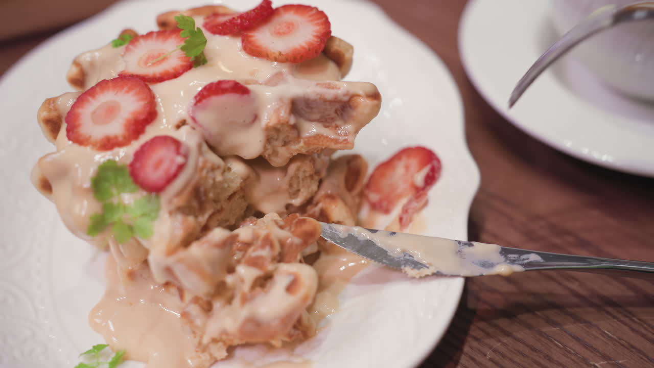 Close-up view of fork picking up fresh strawberry slice from creamy waffle dessert as knife rests on plate. Scene captures detailed textures of pastry on white ceramic dish atop brown wooden table