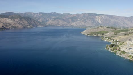 Drone-flying-over-houses-towards-lake-and-mountains