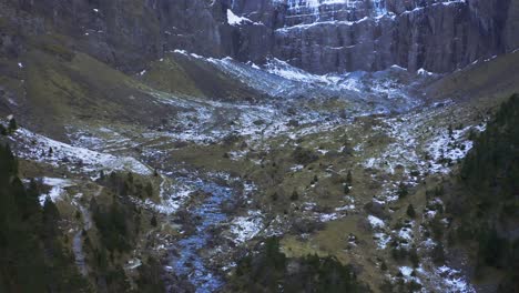 Disparo-De-Un-Dron-De-Un-Acantilado-De-Montaña-Con-Nieve-En-La-Parte-Superior-Que-Se-Desplaza-Hacia-Abajo,-Revelando-Una-Pequeña-Fuente-De-Agua-Del-Río