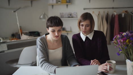 Zwei-Geschäftsfrauen-Arbeiten-An-Einem-Notebook-Und-Hängende-Kleidung-Im-Hintergrund