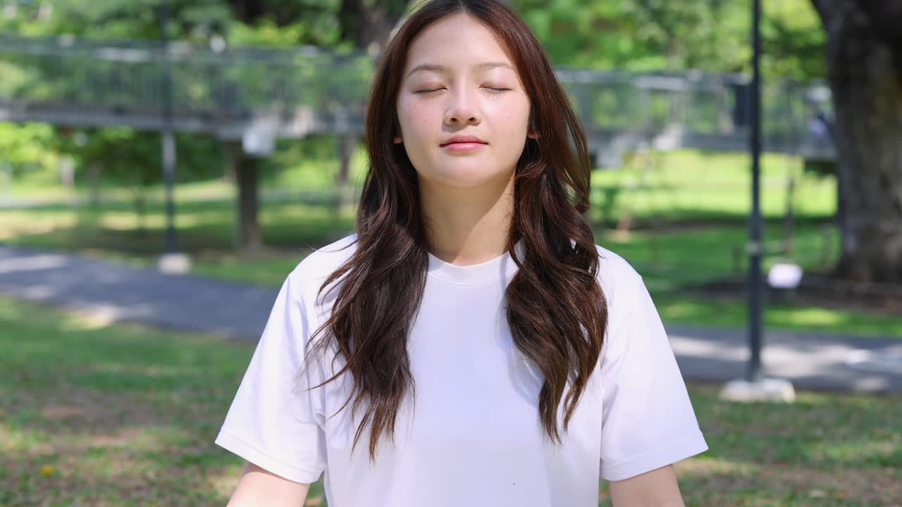 Young Asian woman practices mindfulness meditation outdoors in a sunny park with slow zoom out