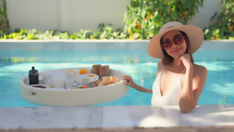 feliz y glamurosa mujer asiática en la piscina con desayuno en un plato flotante