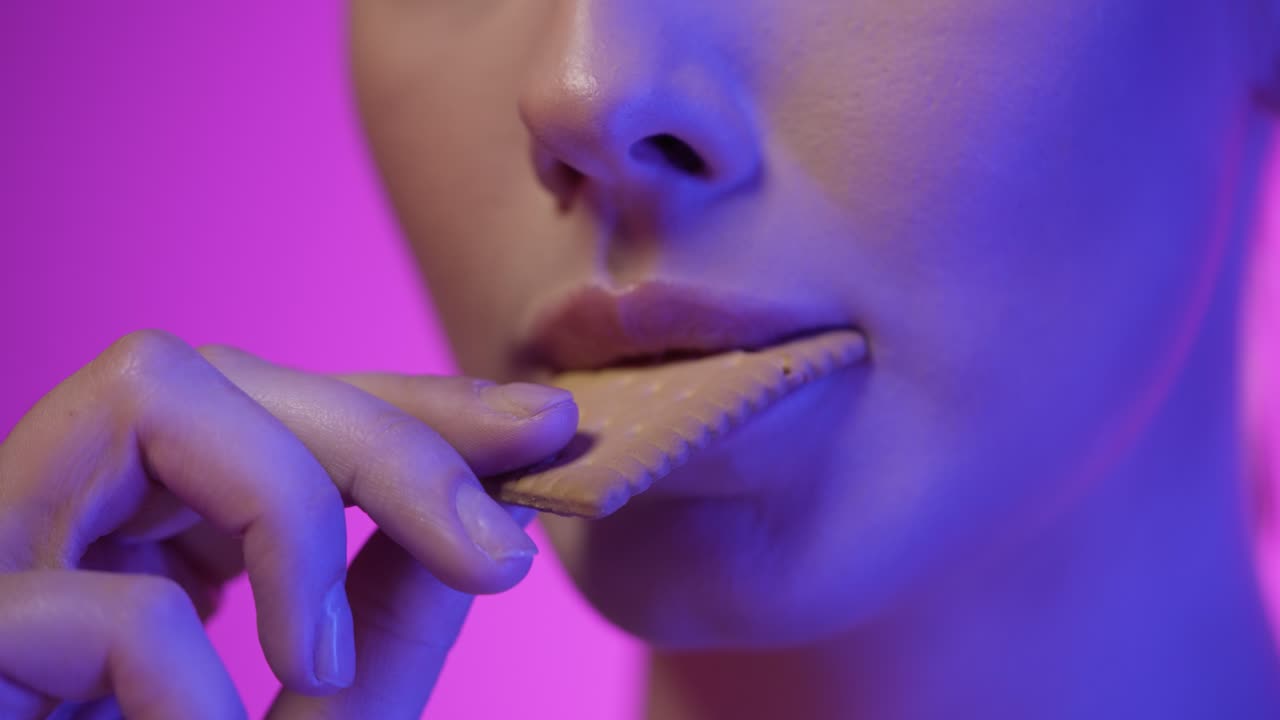 Close up shot of a young pretty woman eating a delicious cookie and enjoying it in front of purple background in slow motion