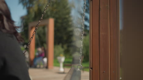 primer plano de una mujer balanceándose al aire libre, su cabello balanceándose suavemente con la brisa, contra un telón de fondo de personas borrosas y vegetación en un parque soleado y brillante