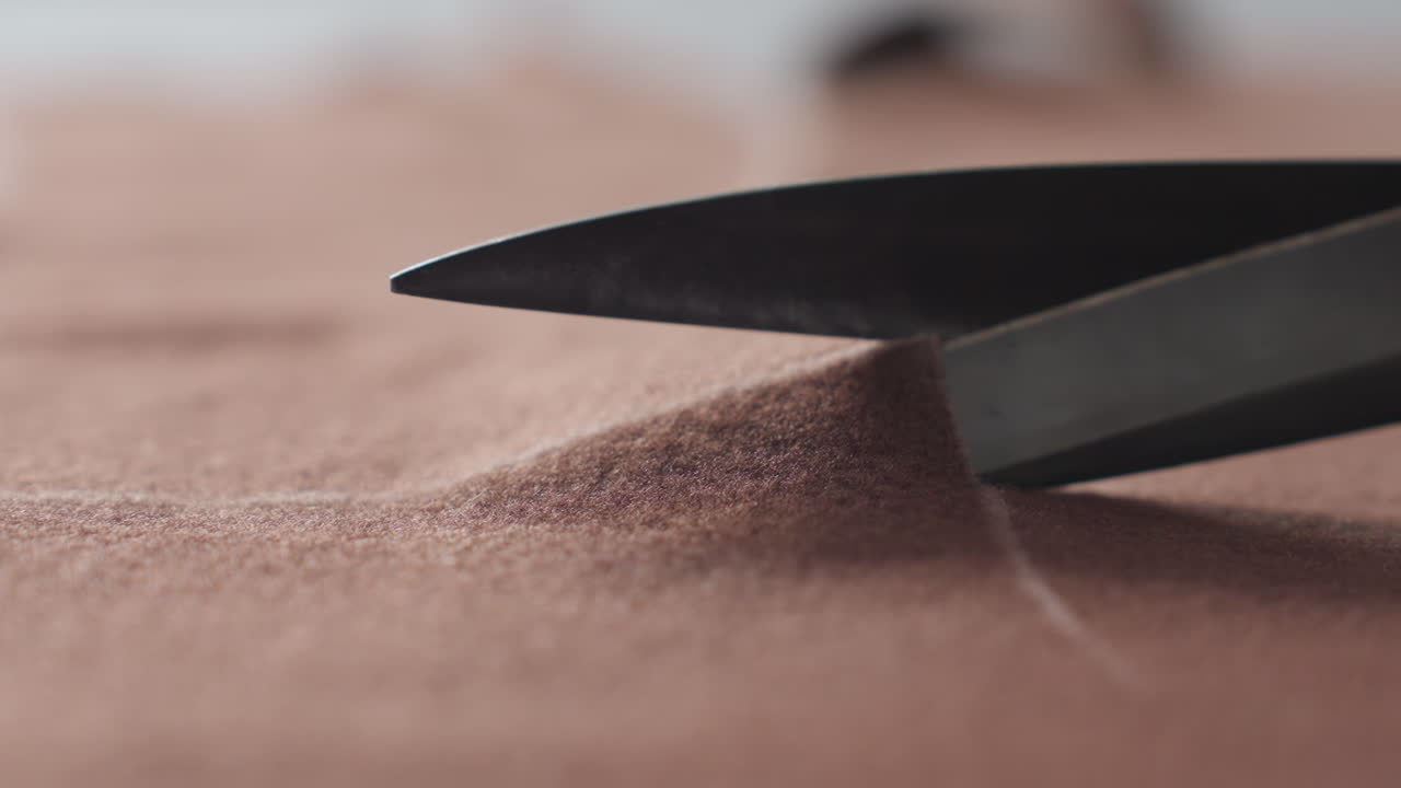 Close up of large metal scissors slicing thick brown fabric along marked white guideline, focused detailed view highlighting precision tailoring process in professional textile or garment workshop