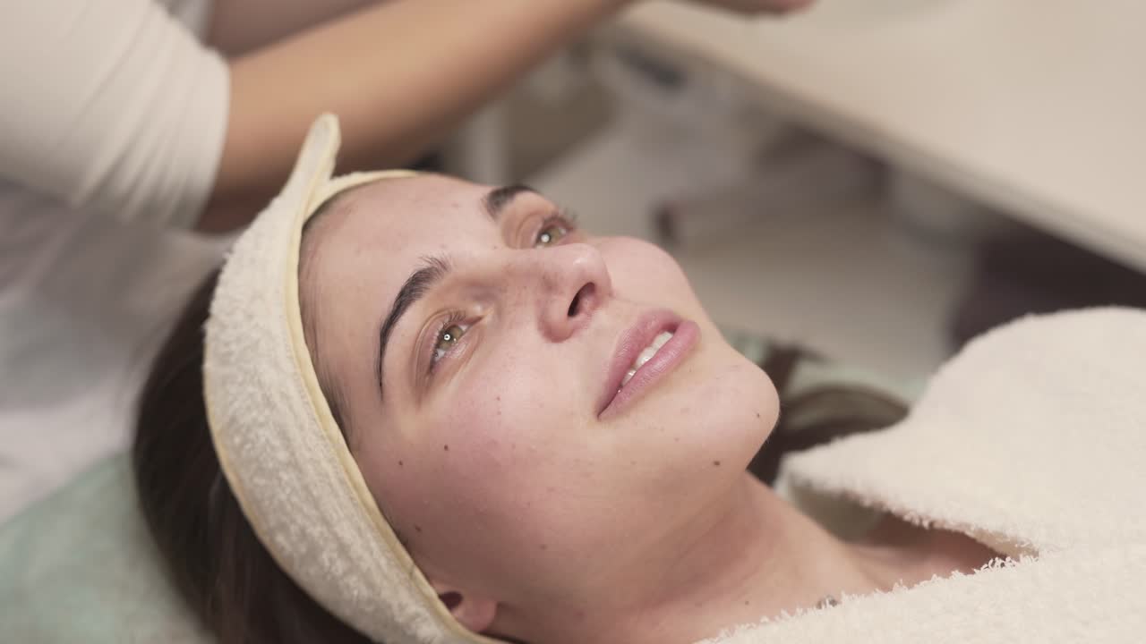 clienta-cliente femenina tendida en el sofá en un hermoso salón de spa, sonriendo y soñando mientras una experta en belleza con bata blanca se limpia la cara con una cinta especial de algodón debajo de los ojos