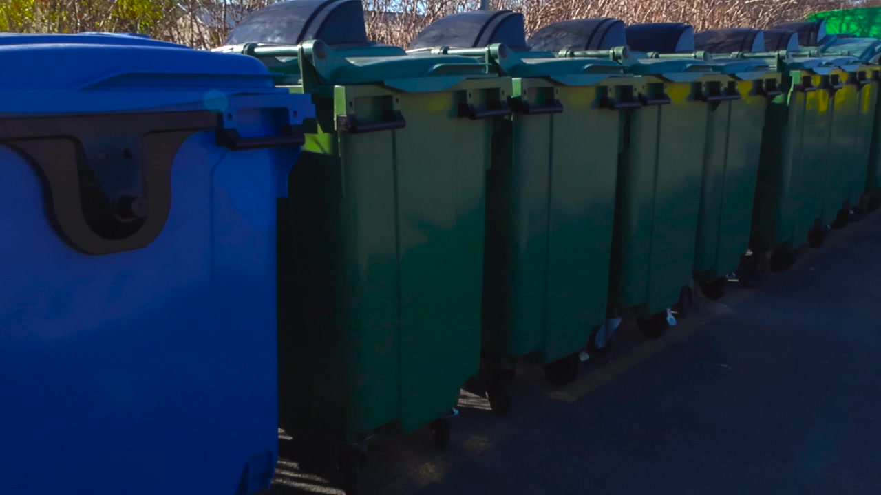 Numerosi bidoni della spazzatura in plastica verdi e blu allineati ordinatamente all'esterno in una giornata di sole, pronti per la raccolta dei rifiuti e il riciclo, evidenziando i concetti di igiene urbana e ambientali