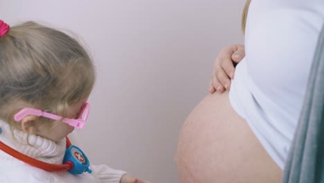 Mädchen-Legt-Zu-Hause-Ein-Spielzeugstethoskop-Auf-Den-Bauch-Der-Schwangeren-Mutter