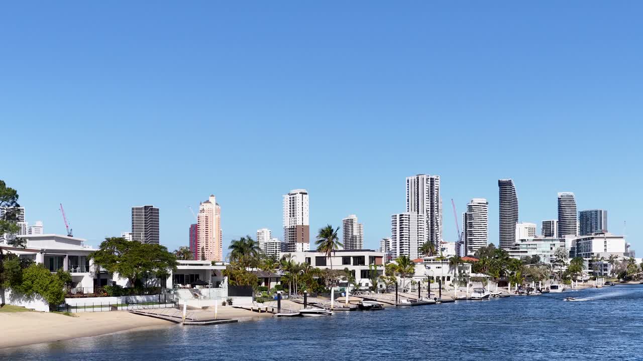 Motorboat glides along river, passing luxury waterfront houses, city skyline, under bright daylight