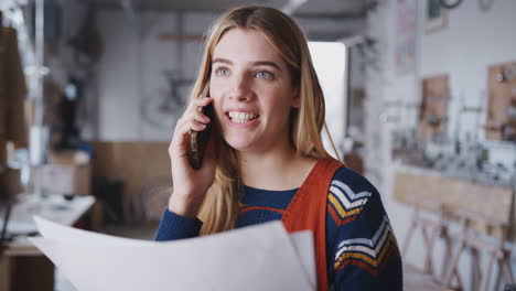 Female-Business-Owner-In-Workshop-For-Building-Bicycles-Making-Call-On-Mobile-Phone