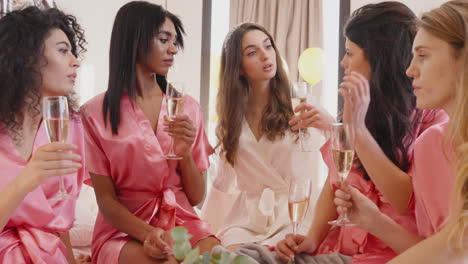 Group-Of-Multiethnic-Female-Friends-And-Bride-Dressed-In-Pink-And-White-Silk-Nightdresses-Talking-While-Holding-Champagne-Glasses