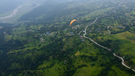 paraglider soars and drops down above terraced fields as raptor bird flies above, high angle aerial overview