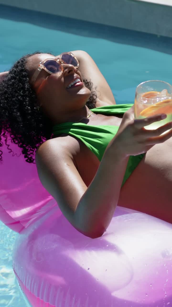 Vertical video: Framing woman in bikini reclining on pink pool float, holding citrus glass, basking