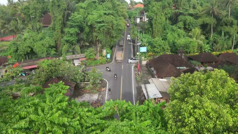 drone shot revealing busy intersection road surrounded by lush dense tropical trees with motorcyclists and cars passing at high speed in traditional balinese district