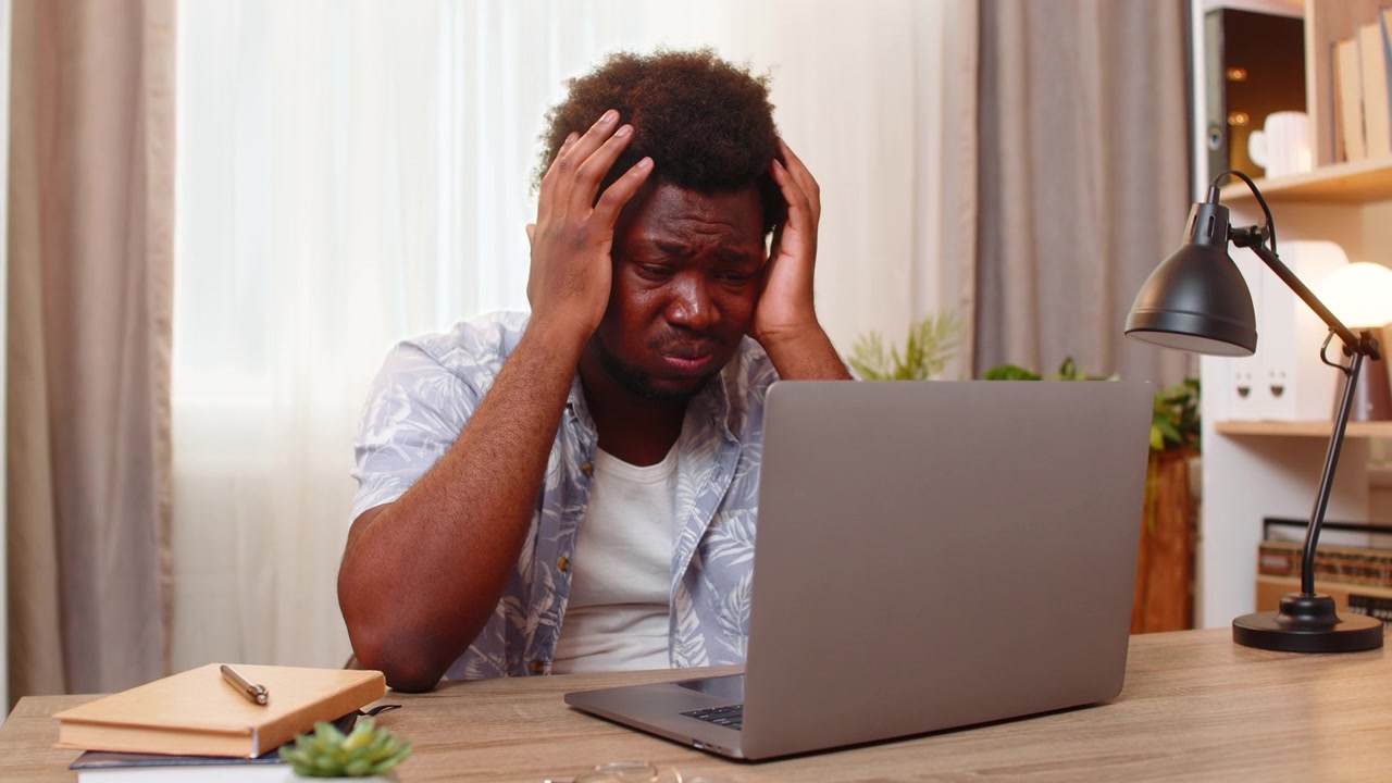 Afroamerikanischer junger Mann am Tisch zu Hause hält schockiert den Kopf, nachdem er schlechte Nachrichten auf dem Laptop-Bildschirm gelesen hat.