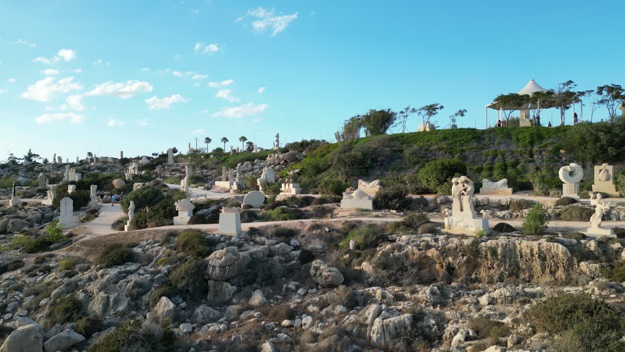 Aerial tracking left of Ayia Napa Sculpture Park with stone statues across rocky cliffside terrain, medium backdrop