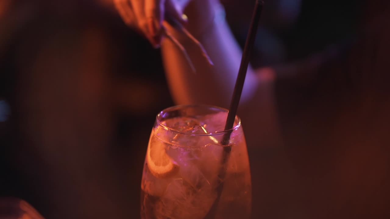 Macro di un barista che usa pinze di metallo per preparare un cocktail ghiacciato con una fetta di agrume e una cannuccia nera in un nightclub o bar dall'atmosfera suggestiva e scarsamente illuminato.