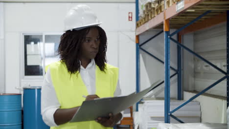 serious african american female worker in helmet counting goods in stock