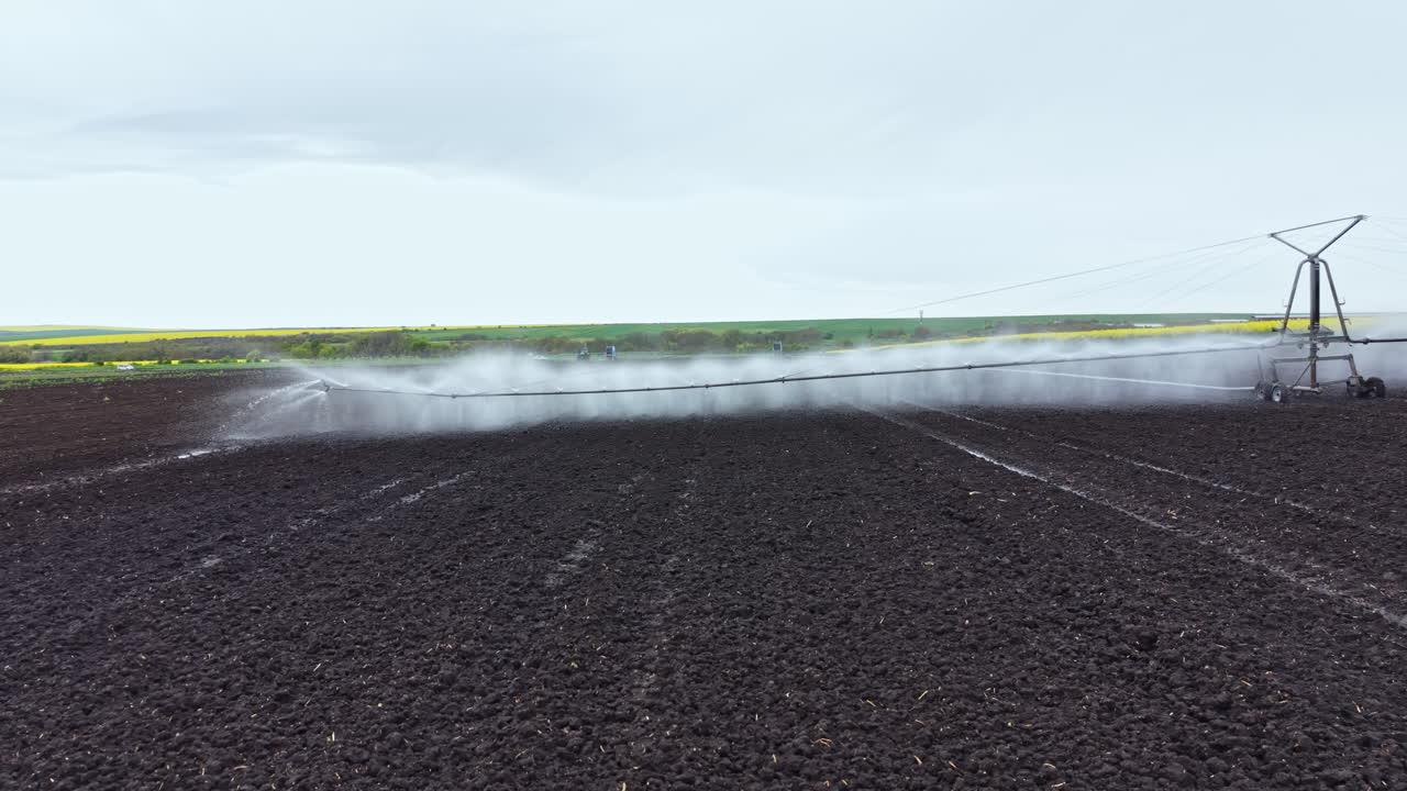 Irrigazione delle colture su un grande campo con un sistema di irrigatori sotto cieli grigi, con campi verdi sullo sfondo, durante la primavera in un'area rurale