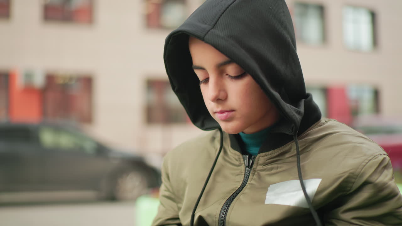 Teen boy in hoodie jacket holding smartphone looks to background with parked cars and residential building behind, appearing curious and observant while seated outdoors in urban setting