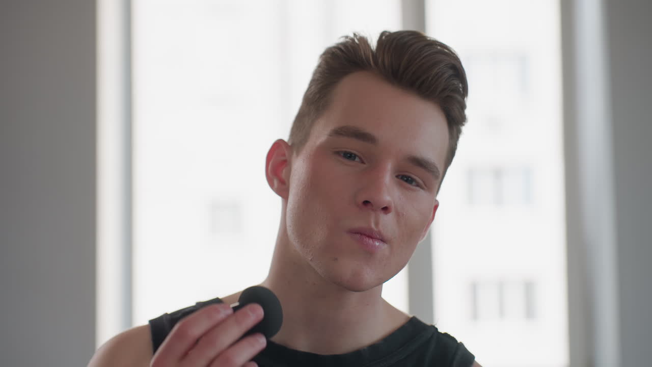 handsome young man in sleeveless signet with neck tattoo sits before bright window mirror, holds black makeup sponge, gracefully dabs powder on cheeks, exudes style and confidence