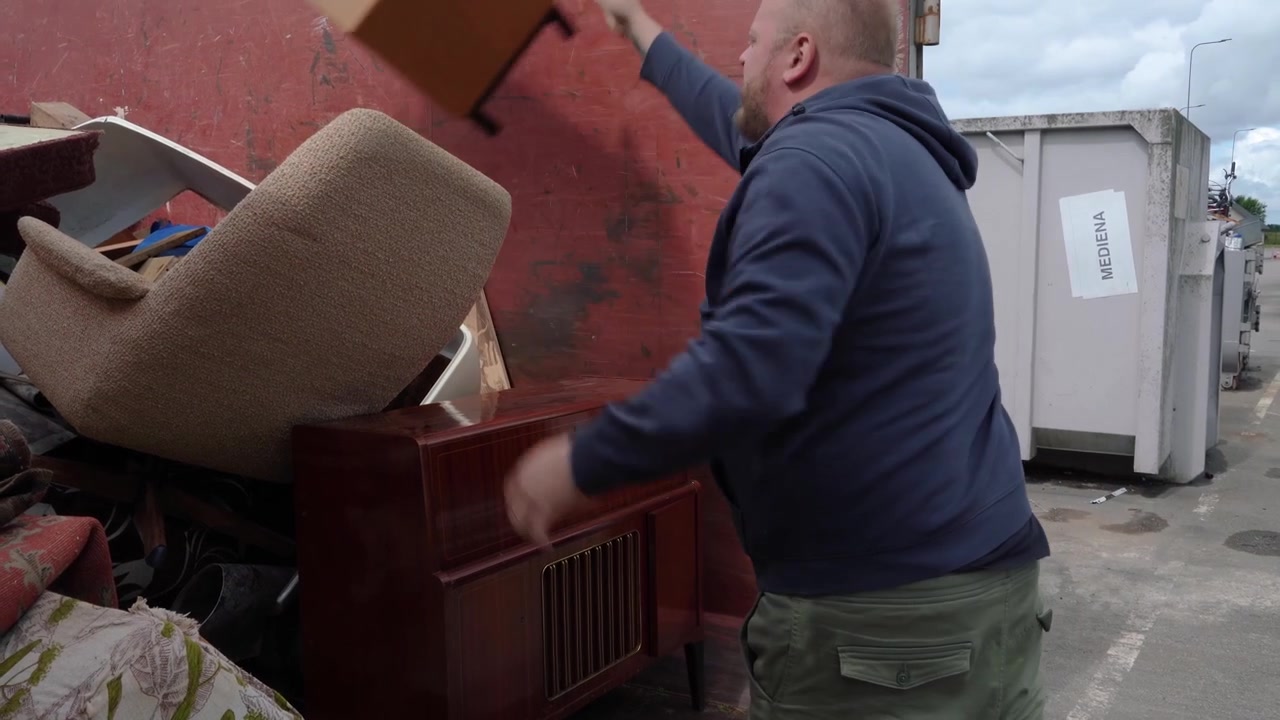 Mann kaster en trehylle på en haug med stort husholdningsavfall ved siden av et gammelt radioskap på en gjenvinningsstasjon