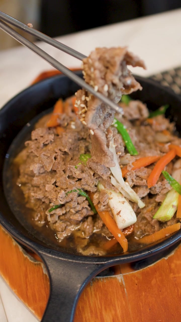 Sumpit mengangkat daging sapi marinasi empuk dari wajan besi cor panas mendesis dengan sayuran dan nasi