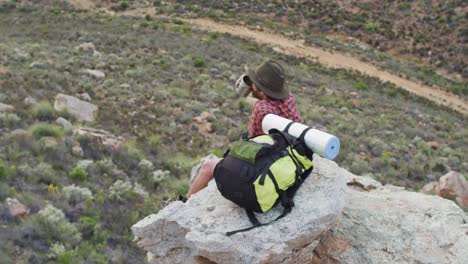 Superviviente-Masculino-Caucásico-Sentado-En-El-Pico-De-Una-Montaña-Rocosa-En-El-Desierto,-Descansando-Y-Bebiendo-Agua