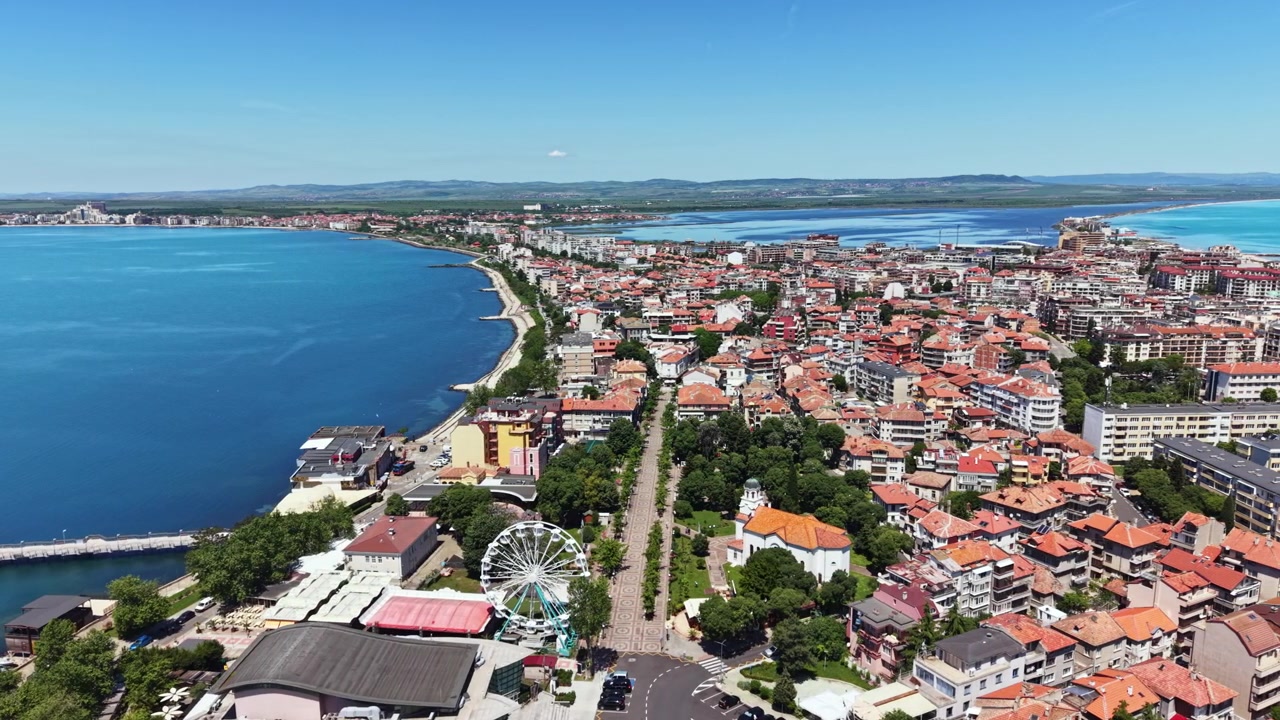 Vista costiera di una città sul mare con edifici, strade e una ruota panoramica in una giornata di sole in una zona costiera.