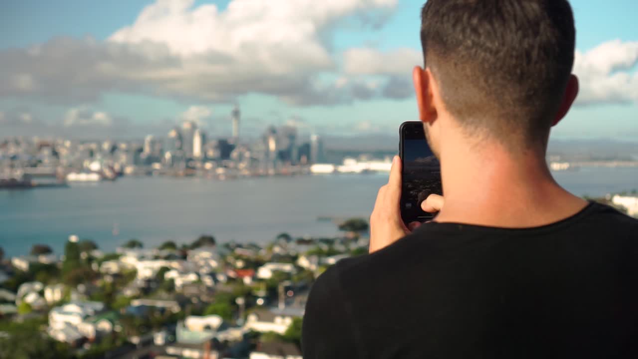 Young caucasian man taking photo with phone of Auckland skyline from Devonport, New Zealand