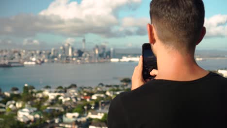 young caucasian man taking photo with phone of auckland skyline from devonport, new zealand