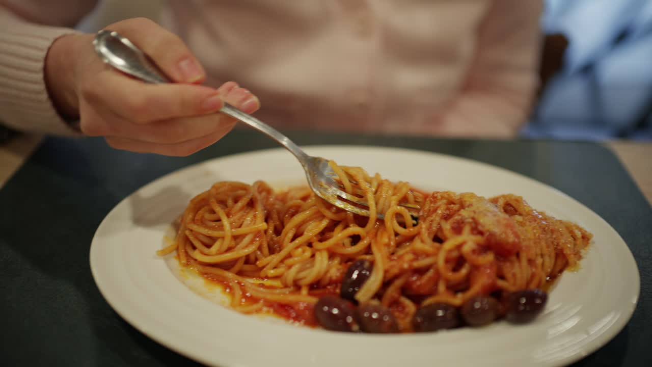 Primo piano di una persona che avvolge spaghetti alla puttanesca con una forchetta, mostrando un ricco sugo di pomodoro, olive e formaggio grattugiato