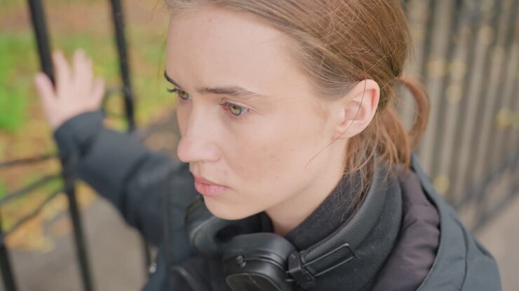 white woman standing by fence, wearing headphones and dark jacket in autumn park, hands resting on iron rail, eyes looking upward with pensive expression, subtle freckles on cheek, cool muted tones