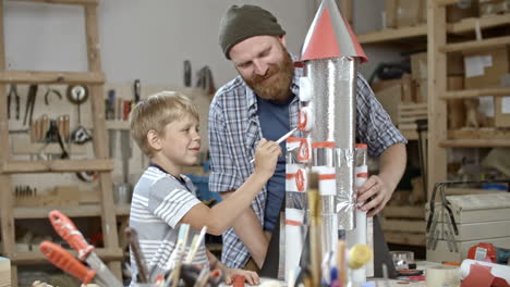 niño pinta un cohete espacial hecho a mano con la ayuda de su padre en el taller de artesanía