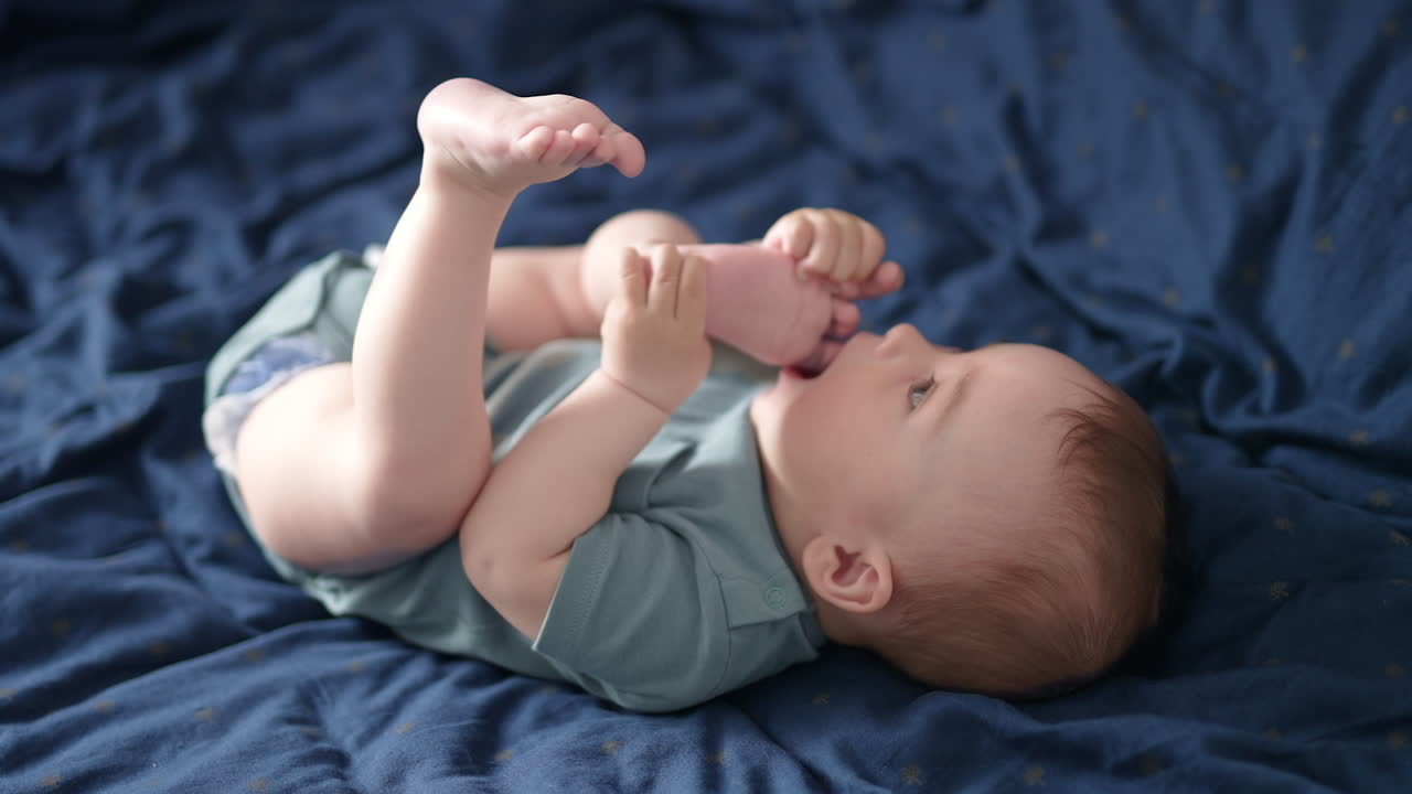 Lovely barefoot child lies on the blue bedclothes. Cute boy pulls his foot to the mouth and chews his toes. Close up.
