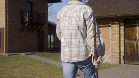 Rear-view-of-a-caucasian-man-in-plaid-shirt-and-cap-holding-an-ax-and-walking-outside-a-country-house