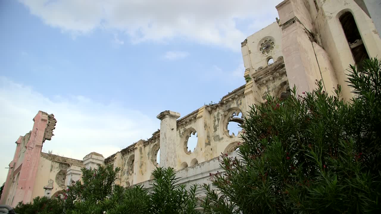 Close-up das ruínas da Catedral de Notre-Dame em Port-au-Prince, Haiti: Fachada gótica desabada com rosáceas circulares, vãos em arco, vestígios de torre rosados, arbustos crescidos e céu parcialmente nublado