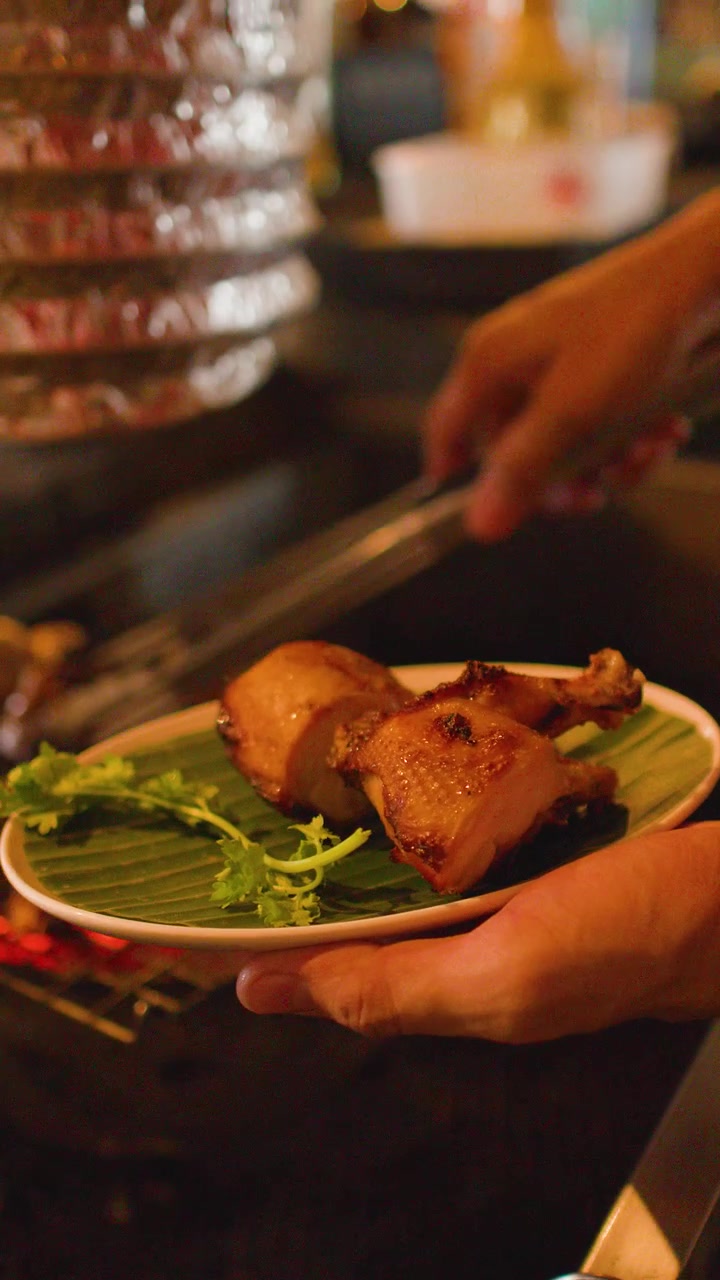 Close-up of hands placing grilled chicken onto a banana leaf plate at a Bangkok market.
