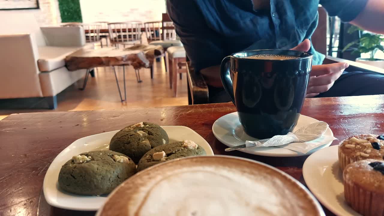 hombre ocupado navegando en su teléfono inteligente mientras está en un café con café humeante, magdalenas y galletas