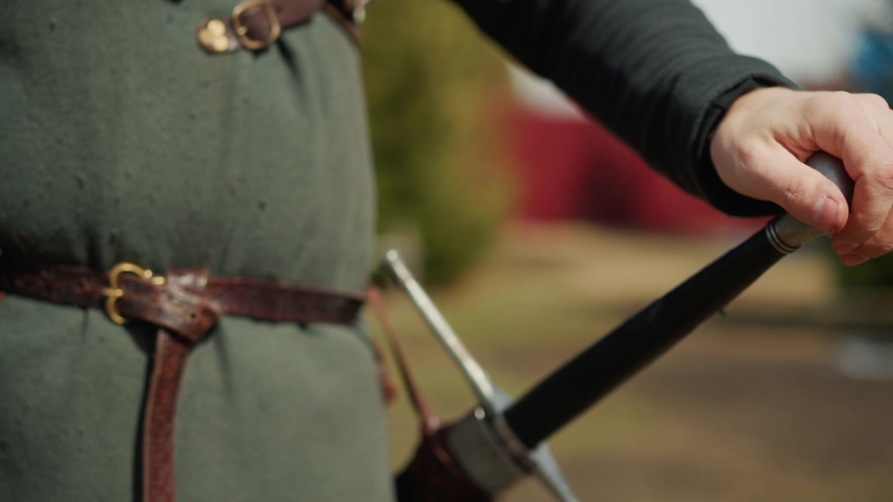 Primo piano di una persona con tunica verde in stile medievale e cintura di pelle che impugna l'elsa della spada vicino al pomolo e alla guardia mentre rivela lentamente la lama dal fodero alla vita, alla luce soffusa del giorno