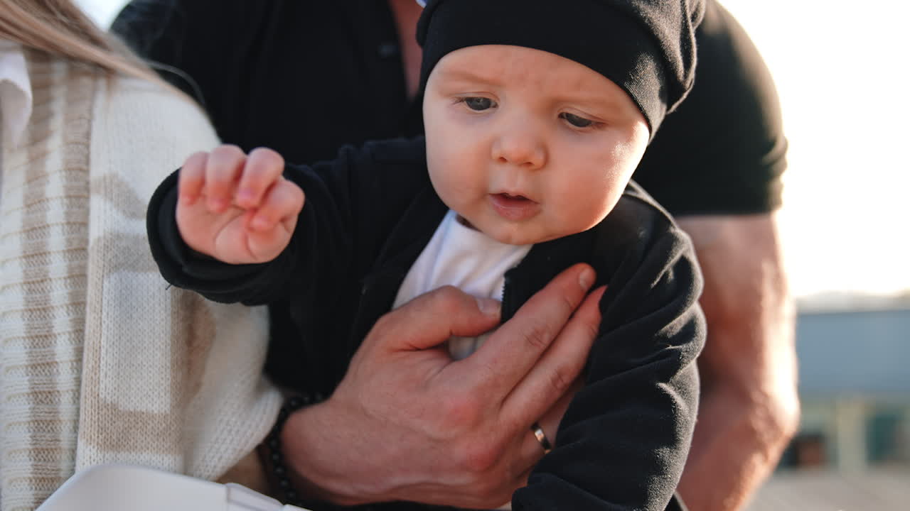 Cute little infant wearing black suit and cap. Baby in hands of parents is interested in the box in front of him. Close up.