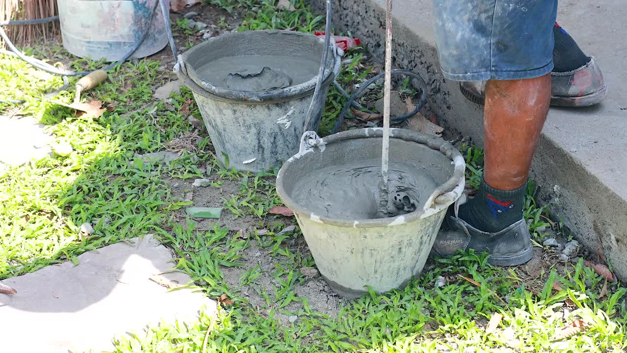 A worker mixes cement in a bucket outdoors under bright sunlight, showcasing construction activity in Phuket, Thailand