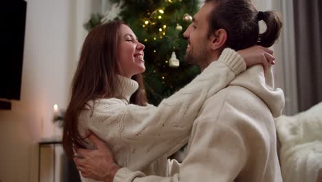 A-brunette-girl-in-a-white-sweater-and-a-guy-and-a-brunette-are-sitting-opposite-each-other-on-the-floor,-looking-at-each-other-and-the-guy-touching-the-girl-on-the-cheek-in-a-cozy-house-decorated-in-the-atmosphere-of-the-New-Year