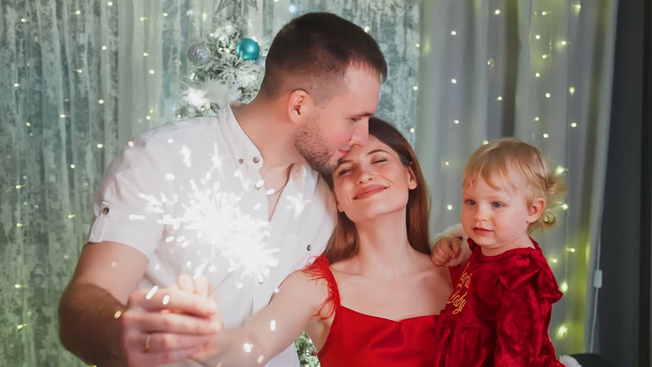 Happy family, mother, father and baby daughter, wearing red clothes, holding sparklers, celebrating Christmas or New Year's Eve near Christmas tree with fairy lights