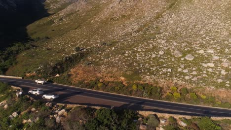 Toma-Aérea-De-Un-Dron-De-Un-Land-Rover-Conduciendo-Por-Un-Paso-De-Montaña-Hacia-Lo-Lejano-Mientras-La-Vista-Se-Extiende-Hacia-Una-Vista-Del-Océano