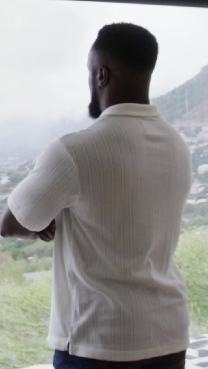 Vertical video: Standing man watching hillside mountain through large window at home, in white polo