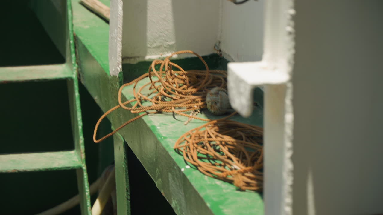 Cuerda naranja enrollada en barandilla verde, primer plano iluminado por el sol de una línea de nailon enredada descansando junto a un mamparo metálico blanco en la cubierta del ferry, almacenamiento práctico y texturas de pintura desgastada, ambiente marítimo industrial