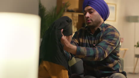 father tying turban onto head of young sikh son with top knot sitting on sofa at home 1